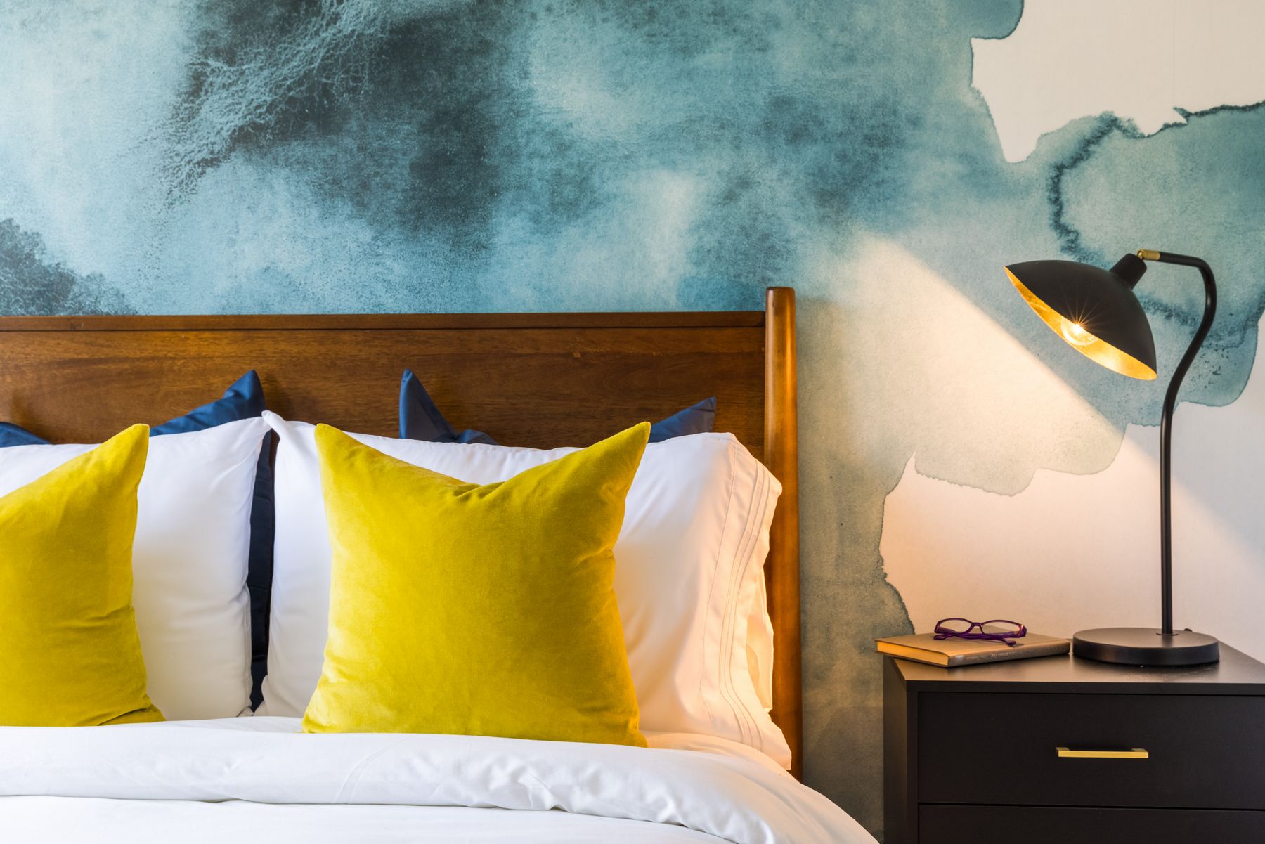 Detail of modern bed corner with yellow, white and dark teal pillows, dark wood night stand with light, book and glasses, teal and white watercolor wall background