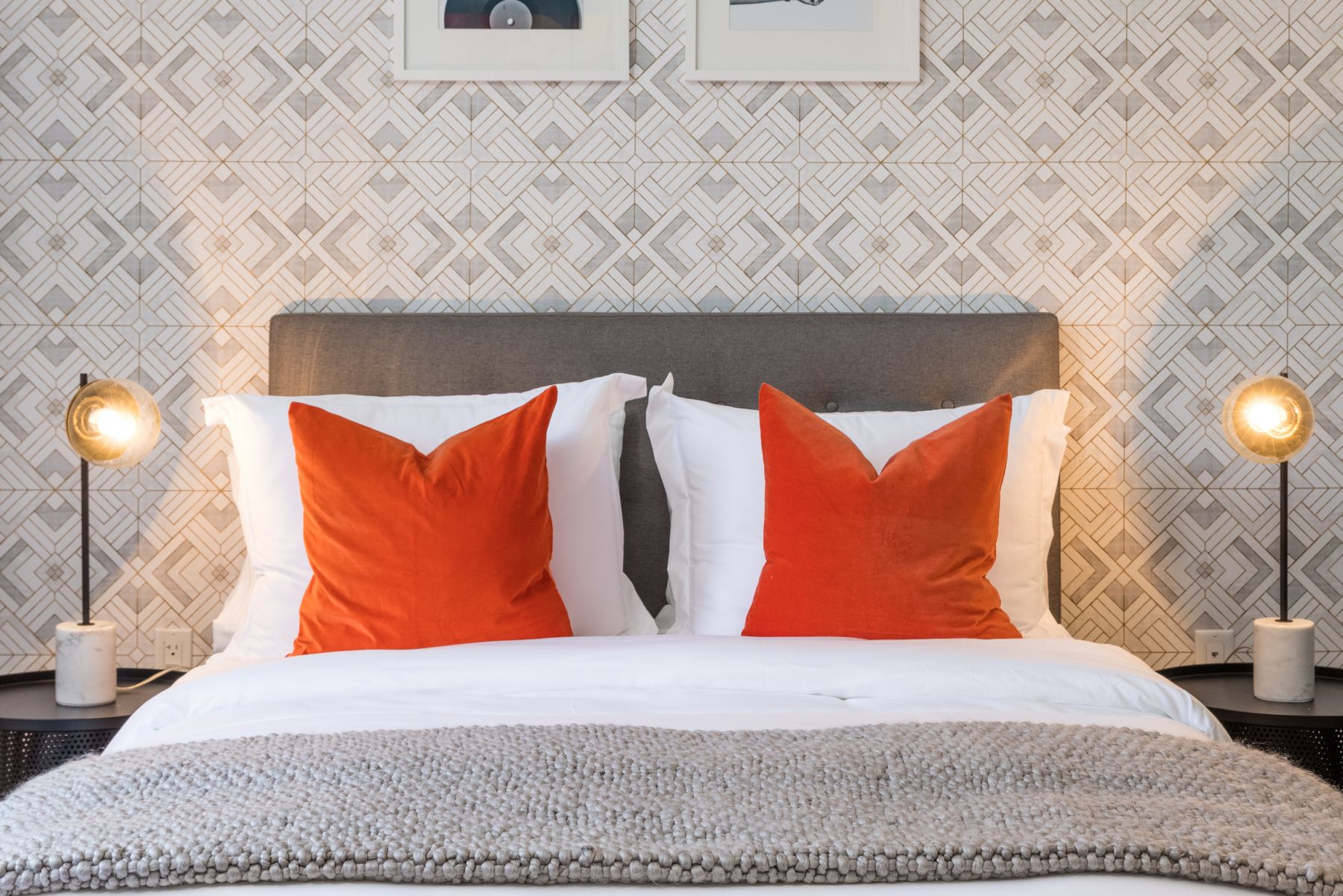 Detail shot of bed with white sheets, duvet and pillows, orange accent pillows, grayish blanket, gray fabric headboard, twin nightstands with light on each side, gray and white patterned wallpaper accent wall behind with frames