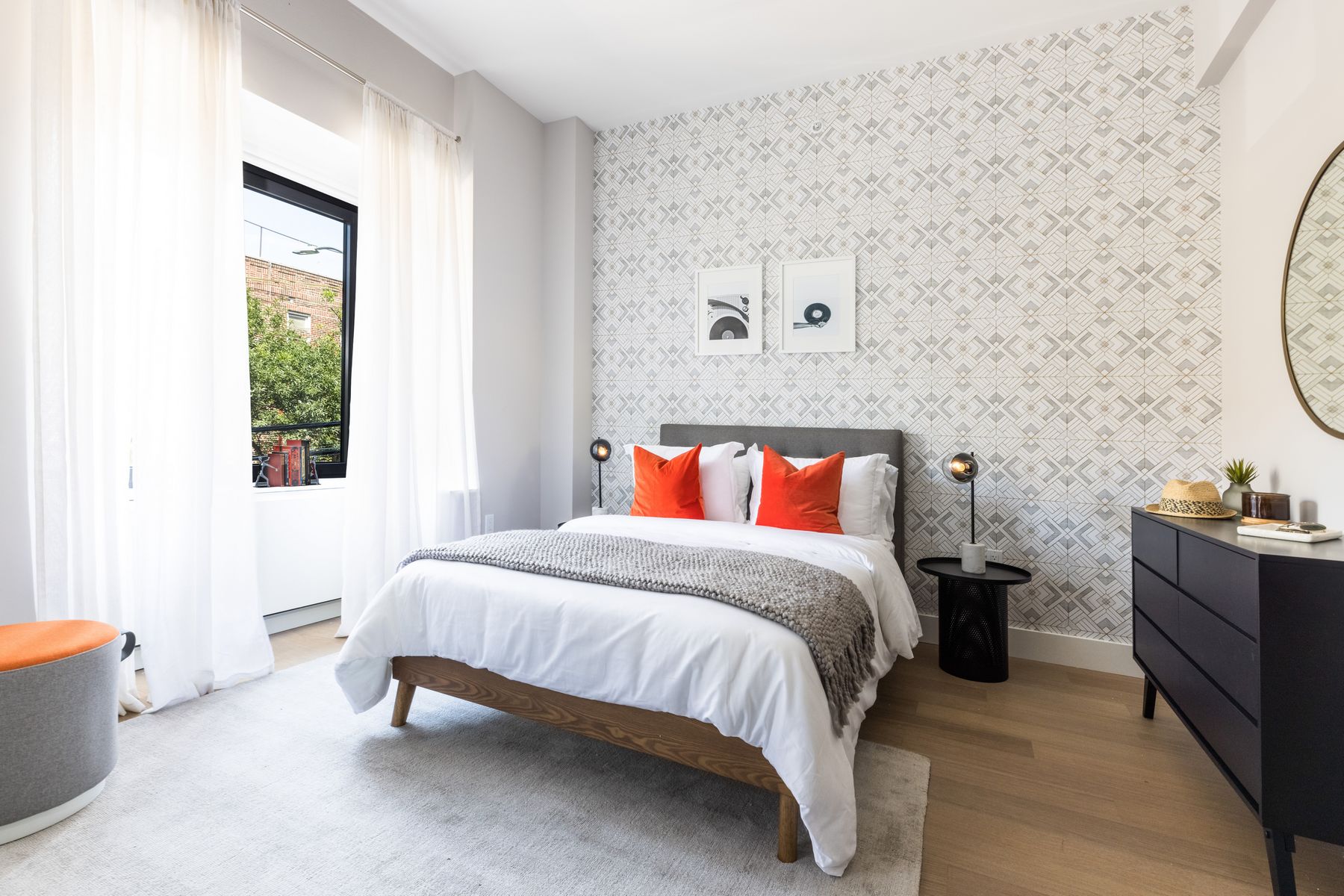 Bright modern bedroom, medium wood fream with grey fabric headboard, orange and white bedding, modern night stand with light, wood floors, dark drawer dresser, white walls with wallpaper patterned accent well behind bed, window on side with white curtains, high ceilings