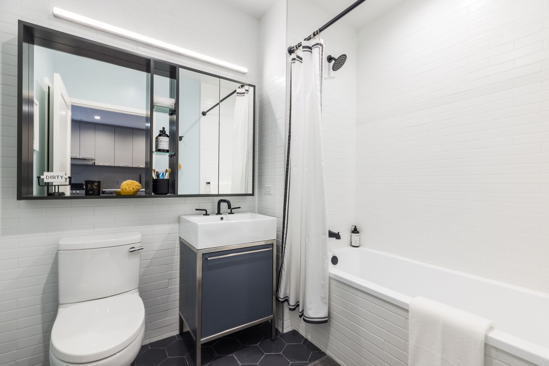 Modern bathroom with fully white tiled tub shower, single vanity with slate blue and silver under sink cabinet, large horizontal glass slider door medicine cabinet above toilet and sink, dark hexagon floor tiles