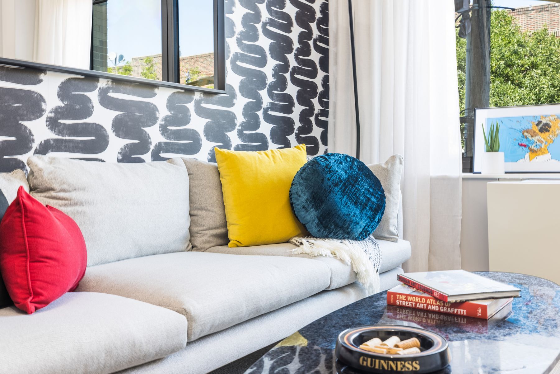 Detail of couch with funky colorful throw pillows, black and white contemporary accent wall, coffee table with books, art, mirror, plant and window with white curtains