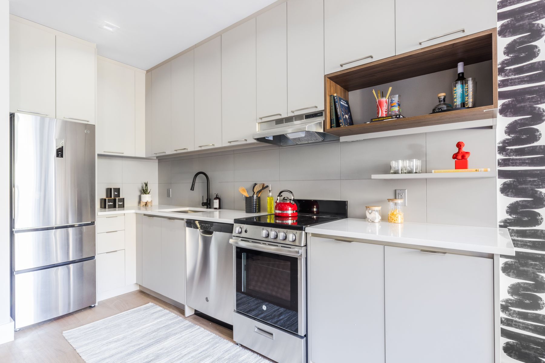 Bright modern galley kitchen with stainless steel fridge, oven/stove and dishwaser, white cabinetry with silver handles and builtin wood shelp, wood flooring