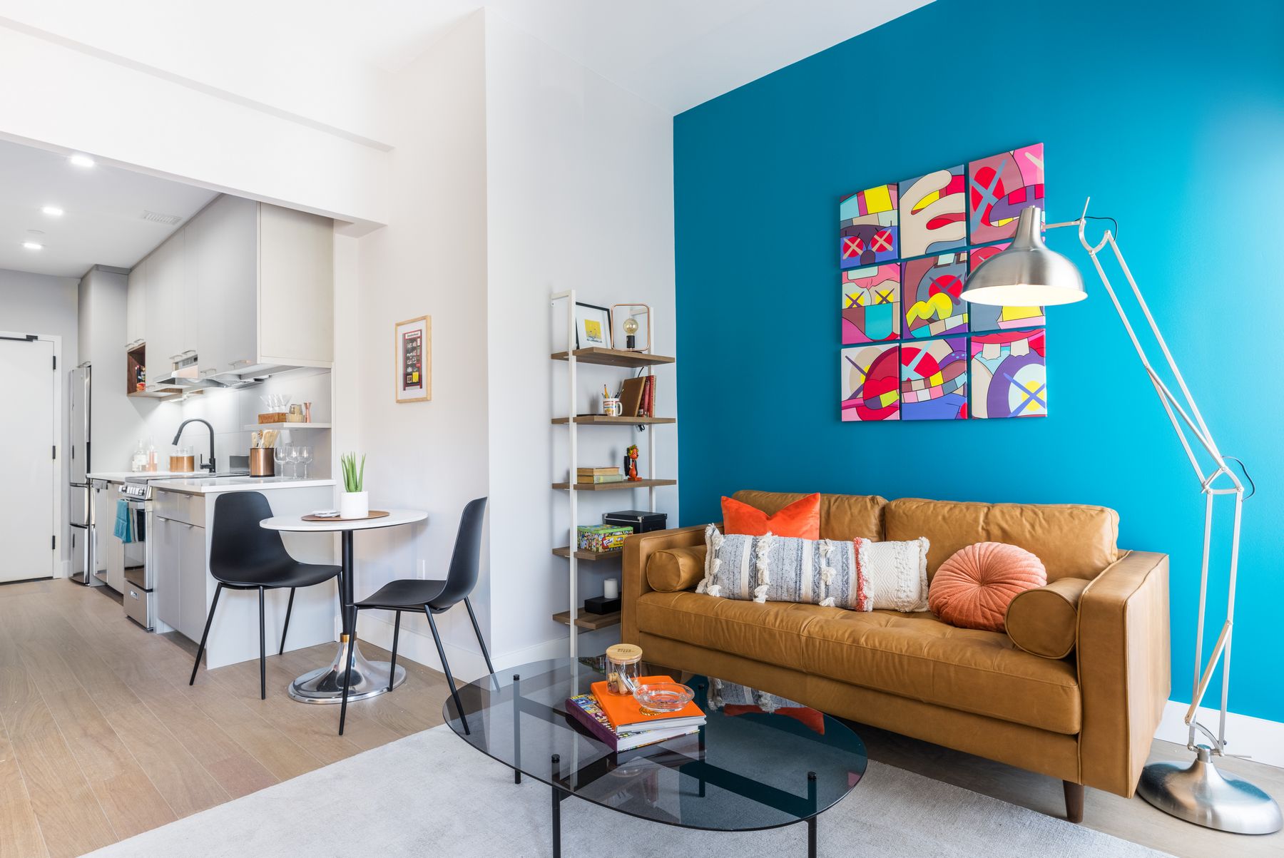 Bright modern open floorplan, camel couch with funky pillows, glass coffee table with books, bookshelf on side, teal accent wall with grid pop part, table for two seating leading to open white kitchen, wood flooring throughout
